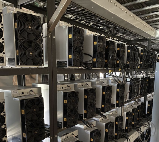 Filas de ASIC para minería de criptomonedas instalados en racks industriales, mostrando ventiladores frontales y cables de alimentación, en una granja de minería a gran escala.
