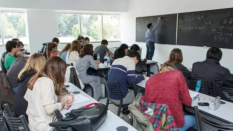 Docente de camisa azul y jeans escribiendo en pizarra negra y alumnos tomando nota