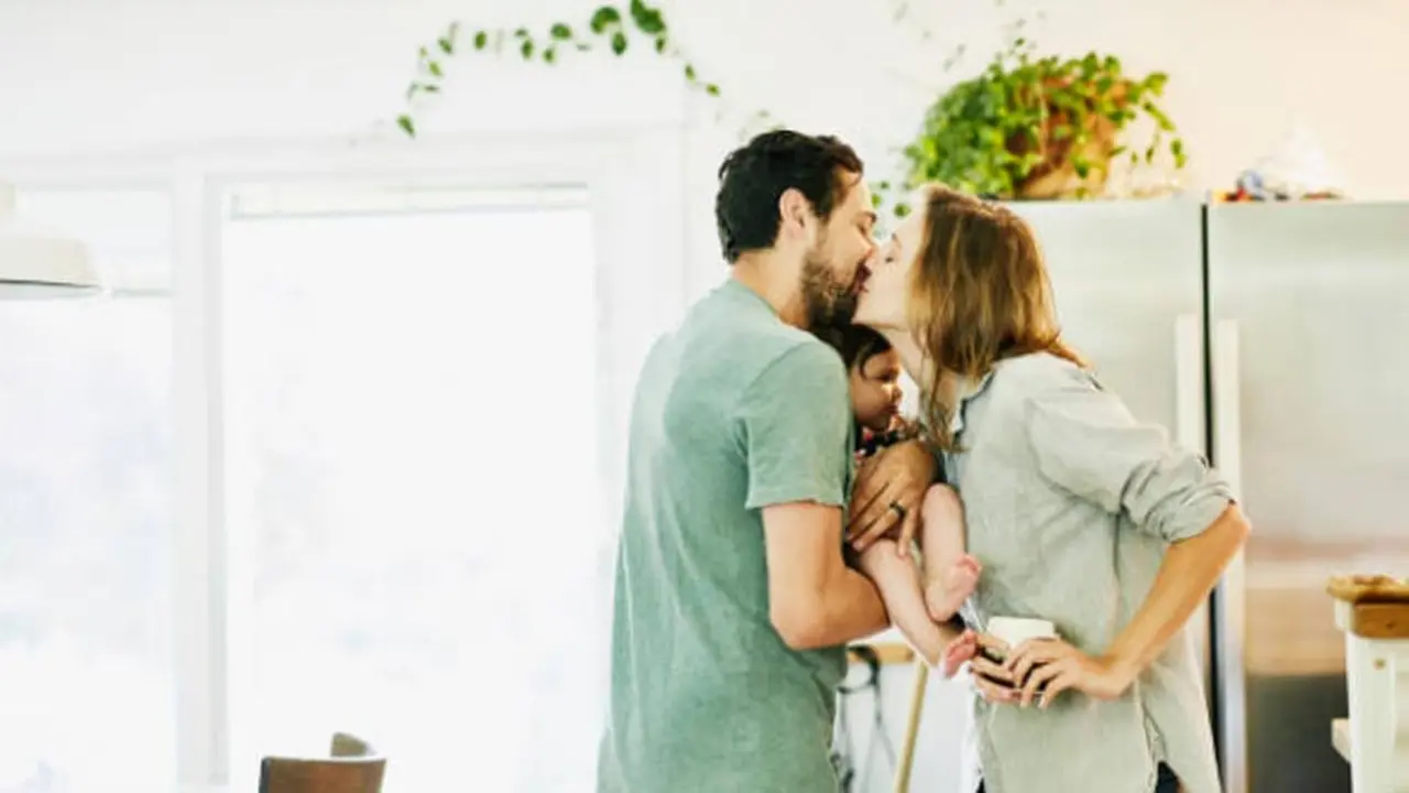 Pareja dandose un beso en una cocina, con el hombre sosteniendo a un bebé. Imagen de archivo.